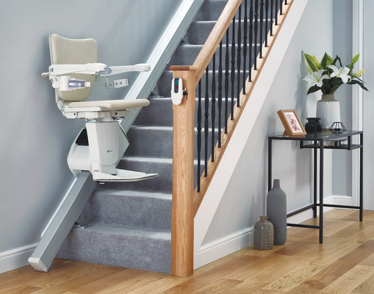 Cream straight stairlift chair parked at the bottom of a grey-carpeted wooden staircase