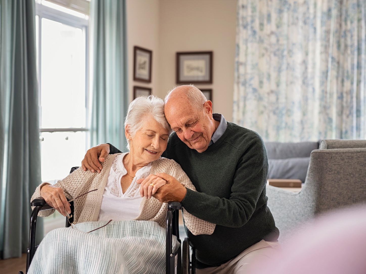 An older man kneeling beside an older woman in a wheelchair, holding her hands and resting his head against hers, in a bright living room
