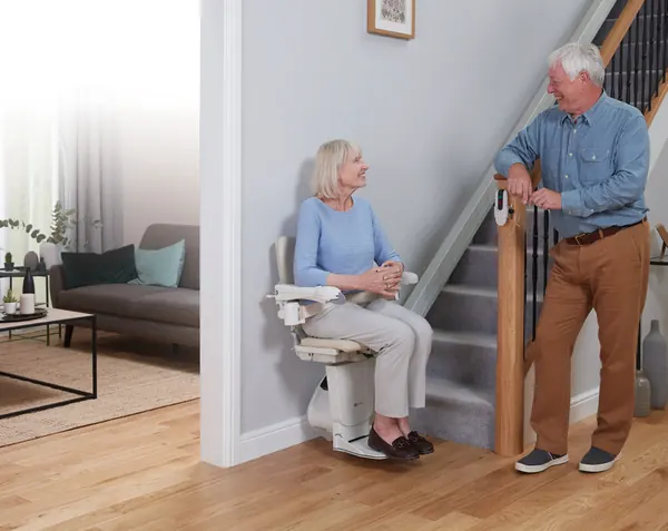 A woman seated on a Handicare 1100 at the foot of the stairs, talking to a man standing beside her