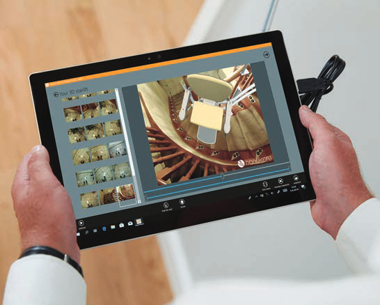 A tablet held in both hands showing a 3D rendering of a Handicare stairlift on a curved staircase, with a seat colour picker down the side