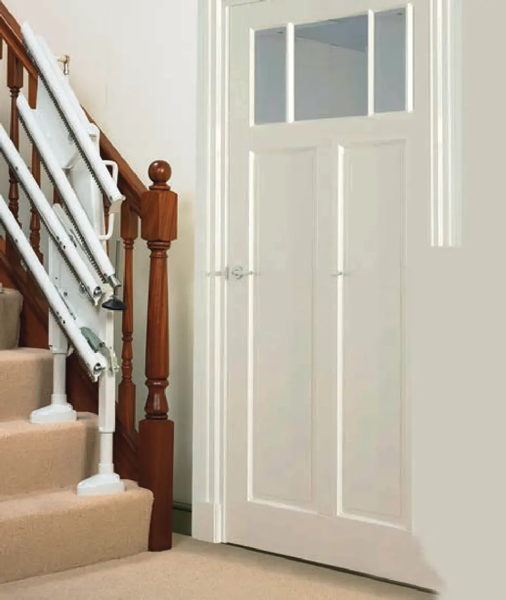 The end of a Handicare stairlift rail beside a white panelled door at the foot of a carpeted staircase