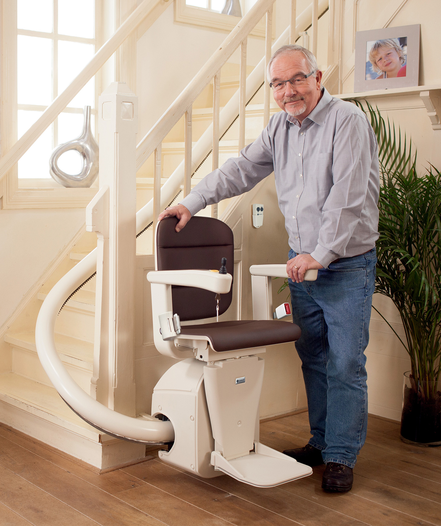 A man standing beside a Handicare Freecurve stairlift parked at the foot of a cream curved rail, resting a hand on the brown seat back