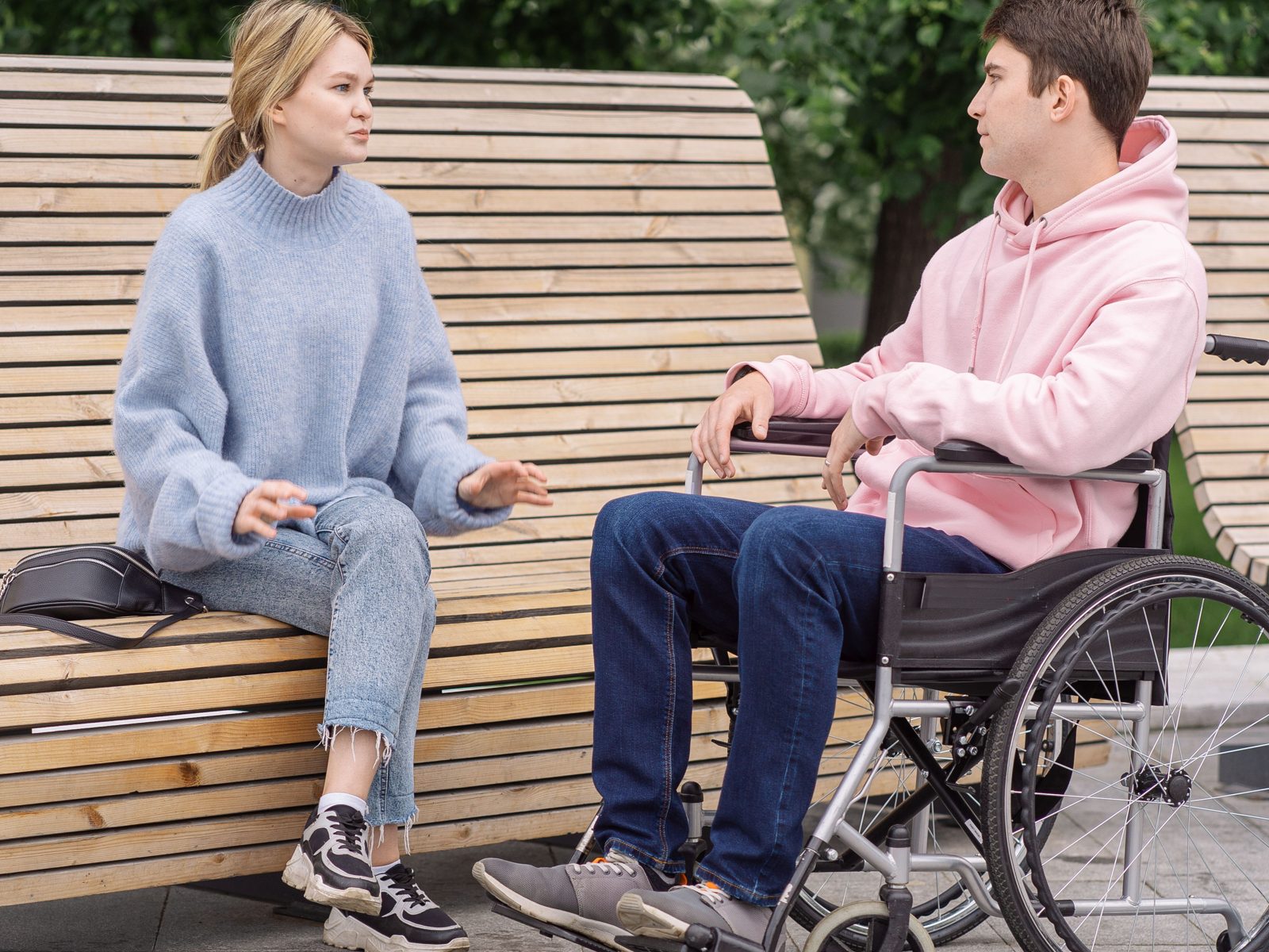 A man seated in a manual wheelchair talking with a woman on a bench in a park