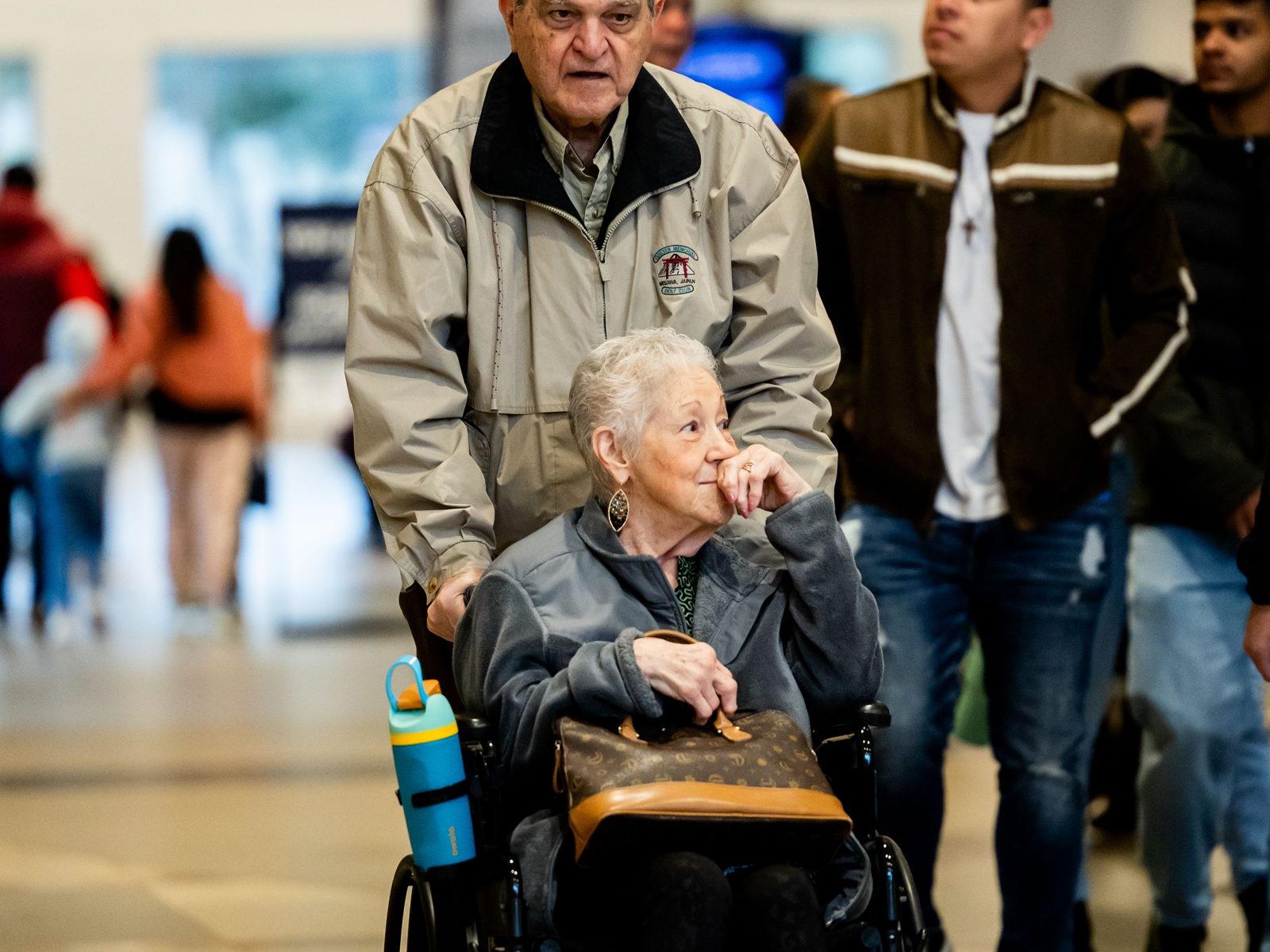 An older man pushing his wife in a wheelchair through a busy public building on an outing