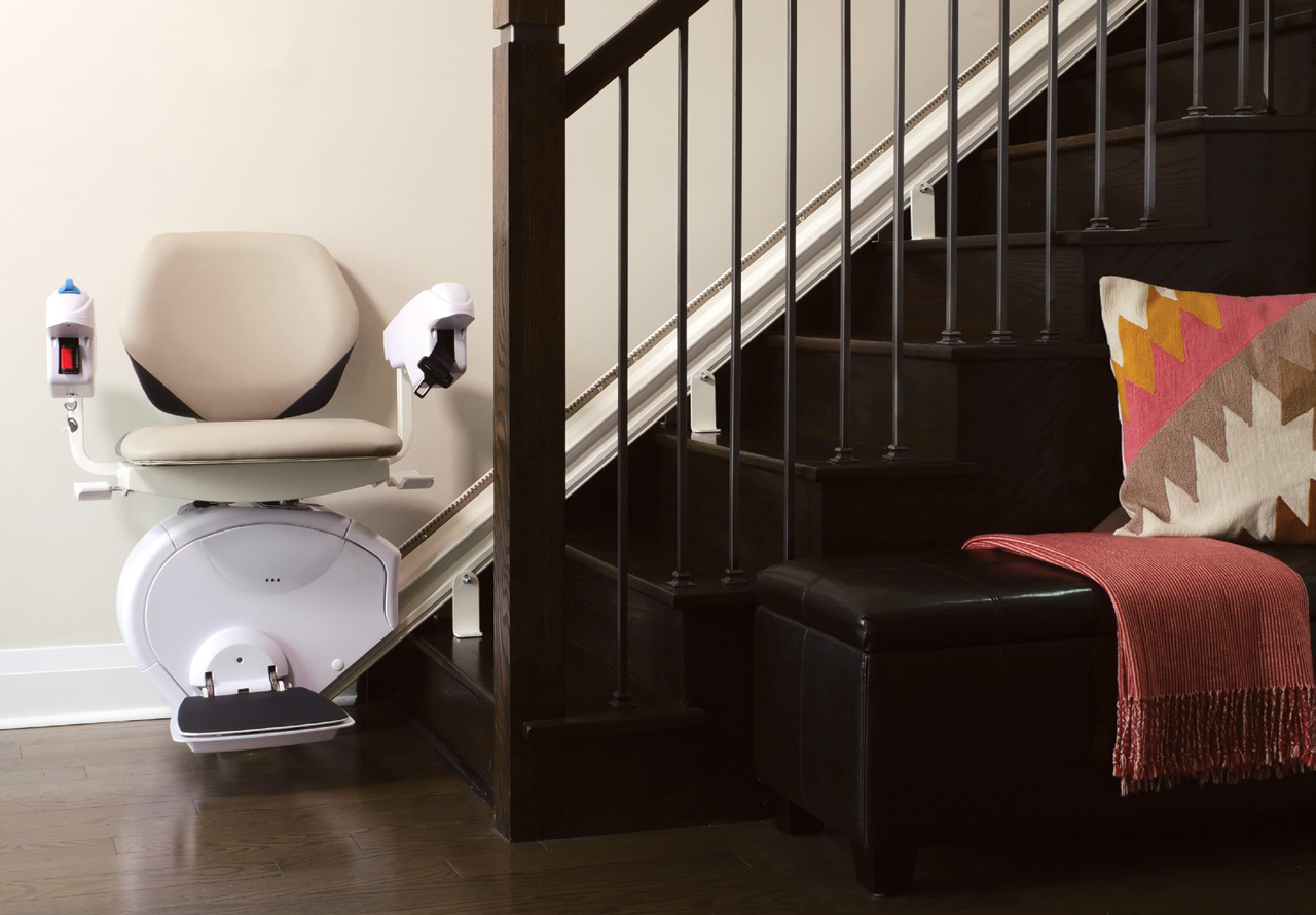 A Savaria K2 Plus stairlift parked at the foot of a dark wooden staircase in a home