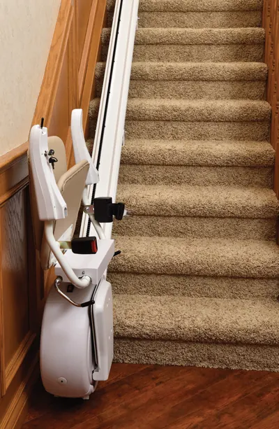 Savaria K2 stairlift folded at the foot of a carpeted staircase, showing the beige seat, armrest and carriage