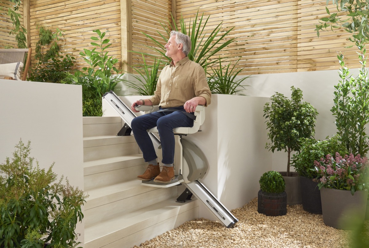 A man seated on an outdoor stairlift beside garden steps